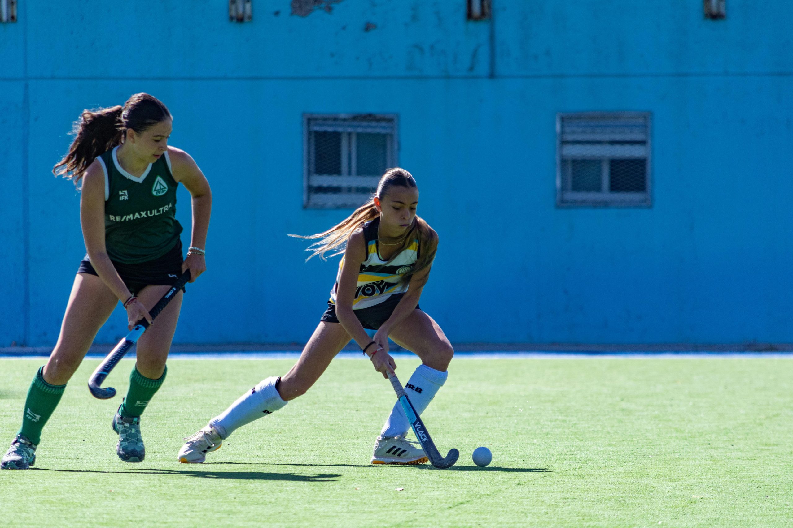 Two young female athletes compete in a lively field hockey match outdoors.